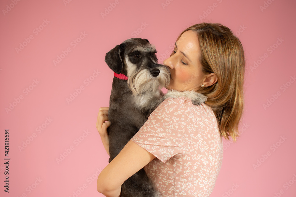Cute young woman hugs her puppy schnauzer dog. Love between owner and dog isolated over pink background. Studio portrait.