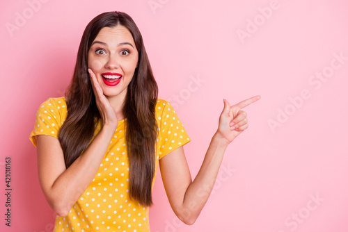 Photo of pretty young lady point empty space hold hand cheek amazed wear yellow polka dot t-shirt isolated on pink color background