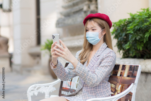 Asian lady who wears suit and red cap holds smartphone to video call with her friend  and sits on bench in park in new normal, healthcare and Coronavirus protection concept. © nut_foto