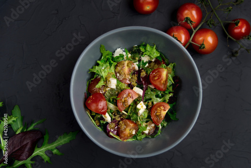 Fresh salad with tomatoes, arugula and cheese cubes on black plate on Dark grey black slate background