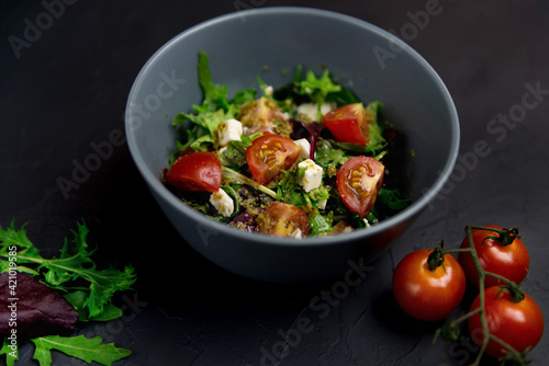Fresh salad with tomatoes, arugula and cheese cubes on black plate on Dark grey black slate background