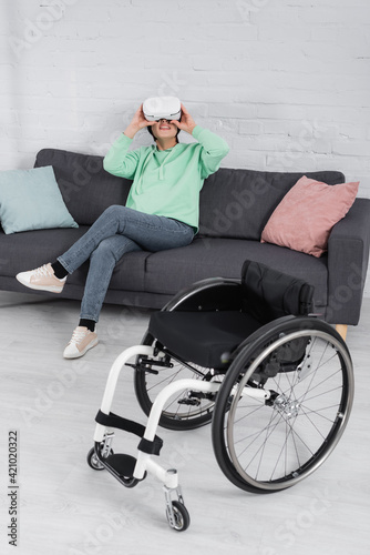 Smiling handicapped woman in vr headset sitting on couch near wheelchair