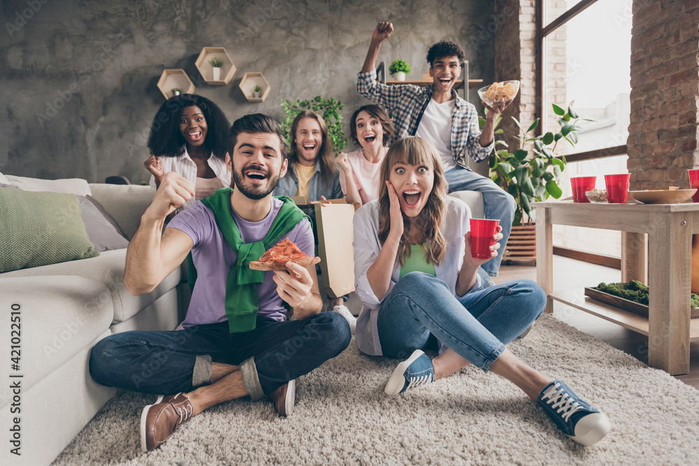 Obraz premium Portrait of nice amazed cheerful friends sitting on carpet having fun eating snack watching cinema laughing in house loft brick style interior indoors