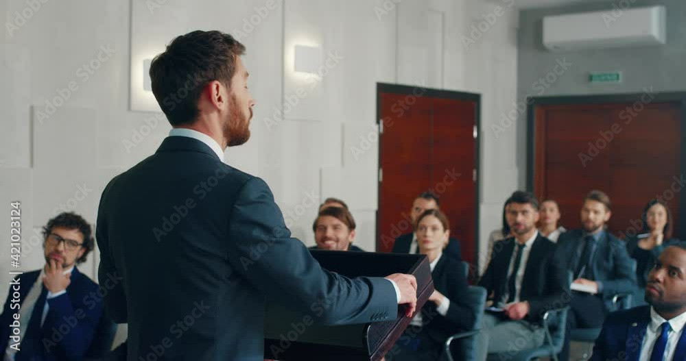 Young successful caucasian entrepreneur speaks in public at business conference standing in front of podium demonstrating presentation.