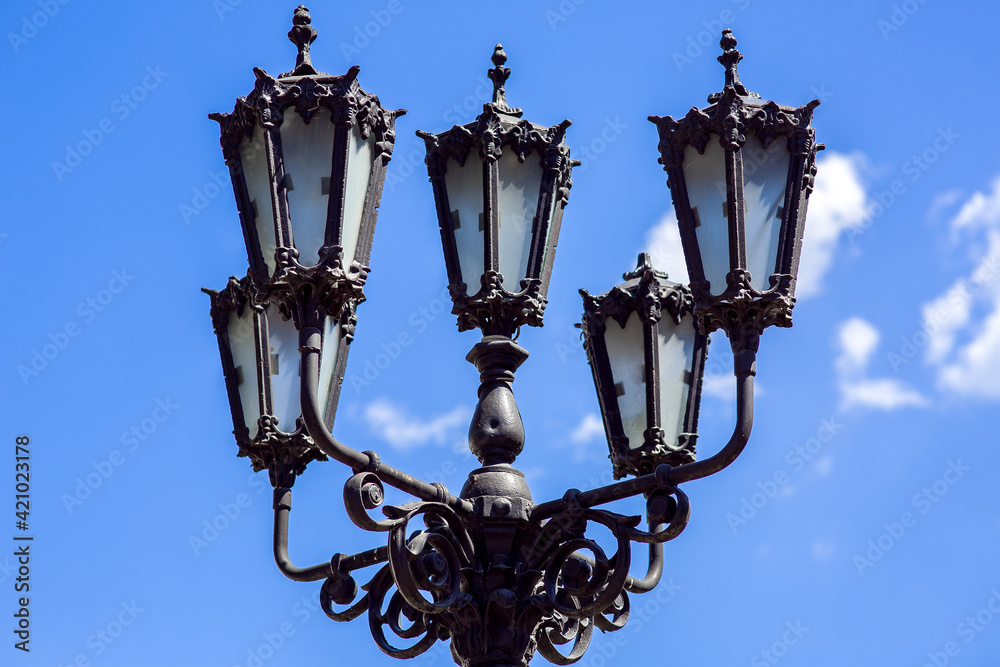 vintage iron street lamp forged from black metal with figured details close-up against a blue sky on a sunny day, nobody.