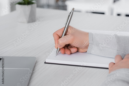 Cropped view of freelancer writing on notebook near laptop on table