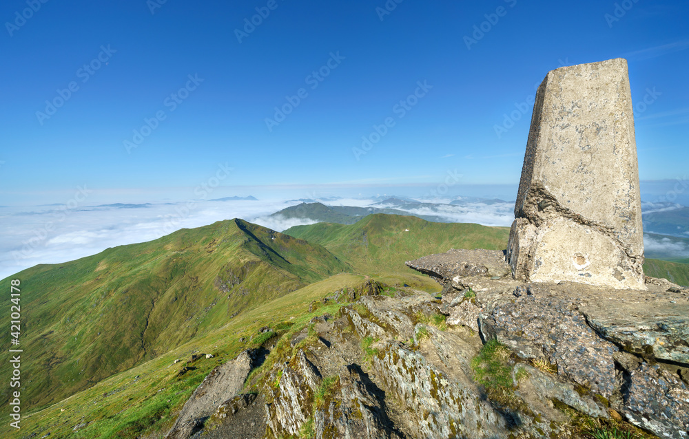 The trig point marker on the mountain summit of Ben Lawers looking out over the summits of Beinn Ghlas and Meall nan Tarmachan with a cloud Inversion covering Loch Tay in the Scottish Highlands, UK.
