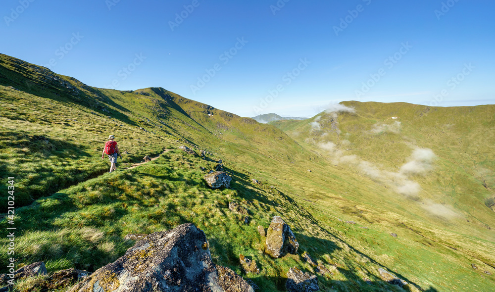 A female hiker and their dog walking towards the mountain summit of Meall Corranaich with the mountain ridge of Ben Lawers and Beinn Ghlas above in the Scottish Highlands, UK landscape.