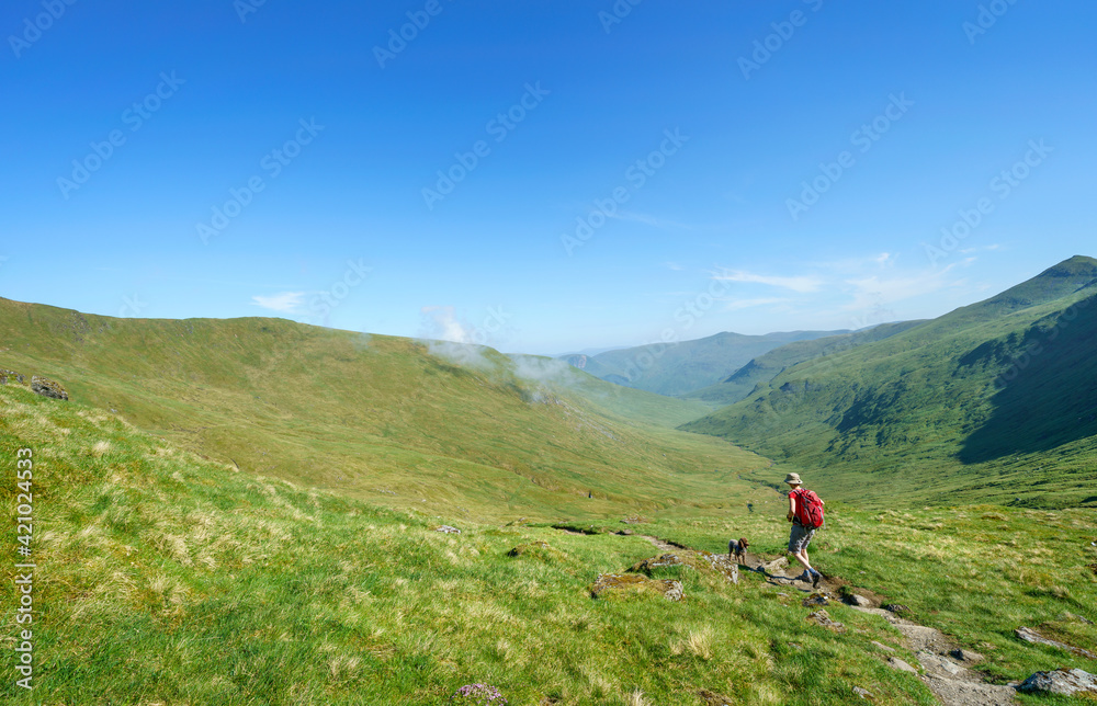 A female hiker walking along a mountain path to the summit of Meall Corranaich with the glen of Allt a Chobhair below and the summit of Carn Gorm in the distance. Scottish Highlands, UK landscape.