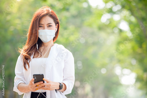 Young asian women Wearing a mask to prevent COVID-19 is using a smartphone on the street on a sunny hot summer day in the park.