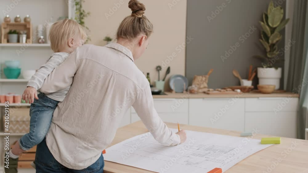 Medium long rear view of woman holding Caucasian toddler son in arms, standing by desk at home, working with blueprint, making adjustments Medium long rear view of woman holding Caucasian toddler son in arms, standing by desk at home, working with blueprint, making adjustments