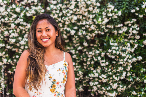 Beautiful portrait of a young tan natural woman with brown and blonde hair smiling wearing a summer dress standing in front of a wall of flowers in San Diego California