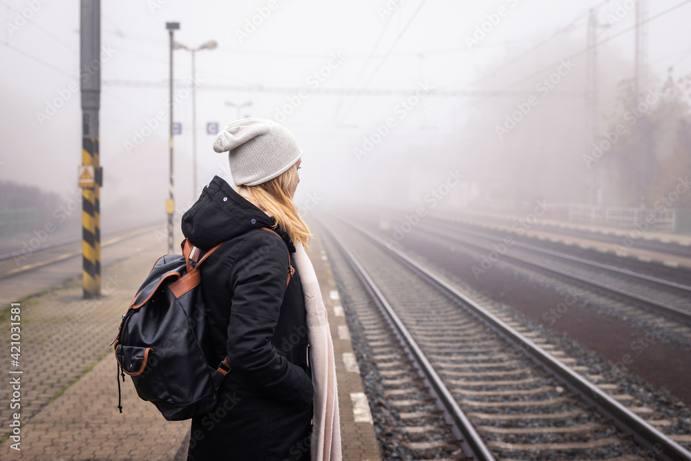 Woman waiting for train at railway station in foggy morning. Female tourist with backpack traveling alone