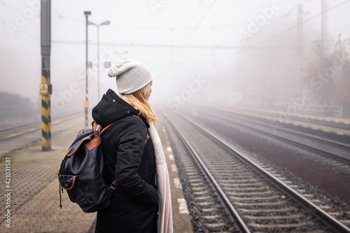 Woman waiting for train at railway station in foggy morning. Female tourist with backpack traveling alone