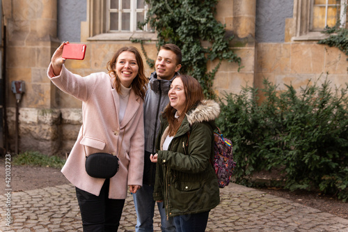 A group of people travel to Europe and record a video blog on their phone. Travel to Hungary, European tour. A group of people - a guy and two girls take a selfie near the old building. © Nona Liuter