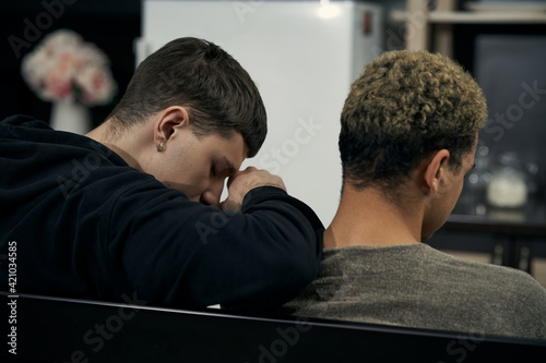 Staged photo illustrates problems and conflicts in gay couple relationships. Moment of showdown: two young men are sitting in the kitchen. Black-haired man's leant on partner's shoulder.