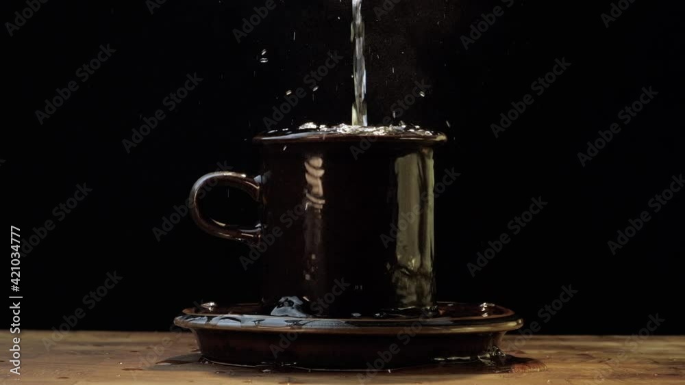 Cup overflows onto the saucer and table. Slow motion. Dark studio