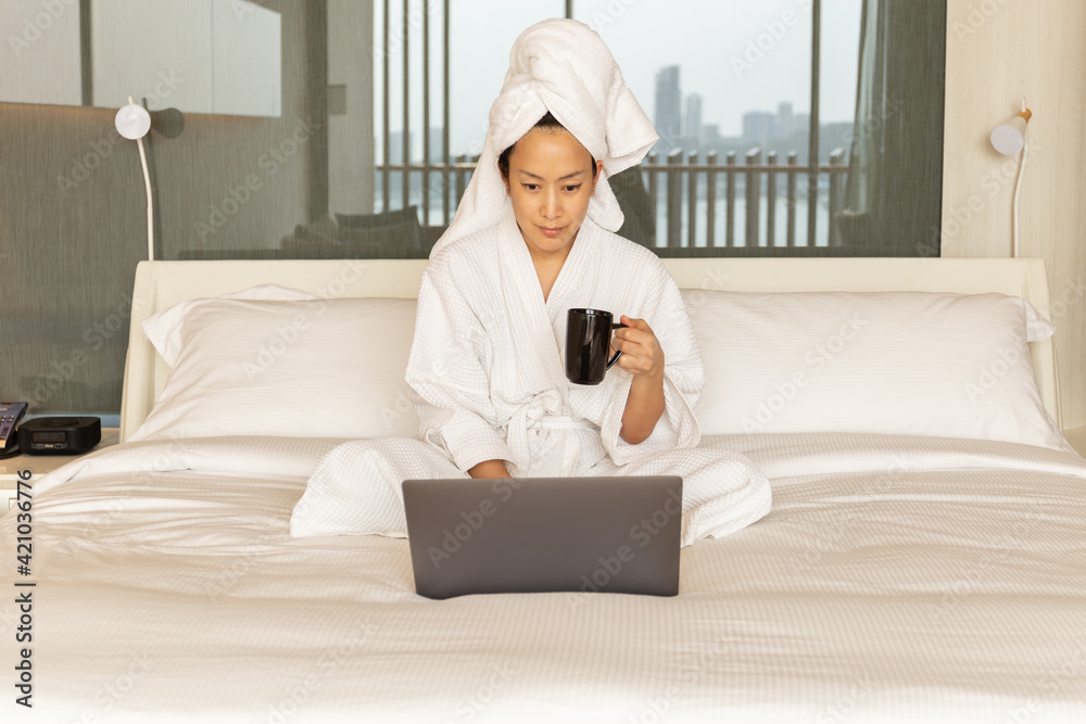 Woman in dressing gown and towel on her head sit on bed hand holding coffee working on laptop. Woman in dressing gown and towel on her head sit on bed hand holding coffee working on laptop.