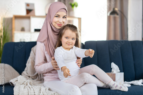 Young smiling Arabian mother with her cute little daughter, sitting on the blue sofa in cozy living room. Pretty child showing hygienic cotton swabs to camera. Hygiene and care concept