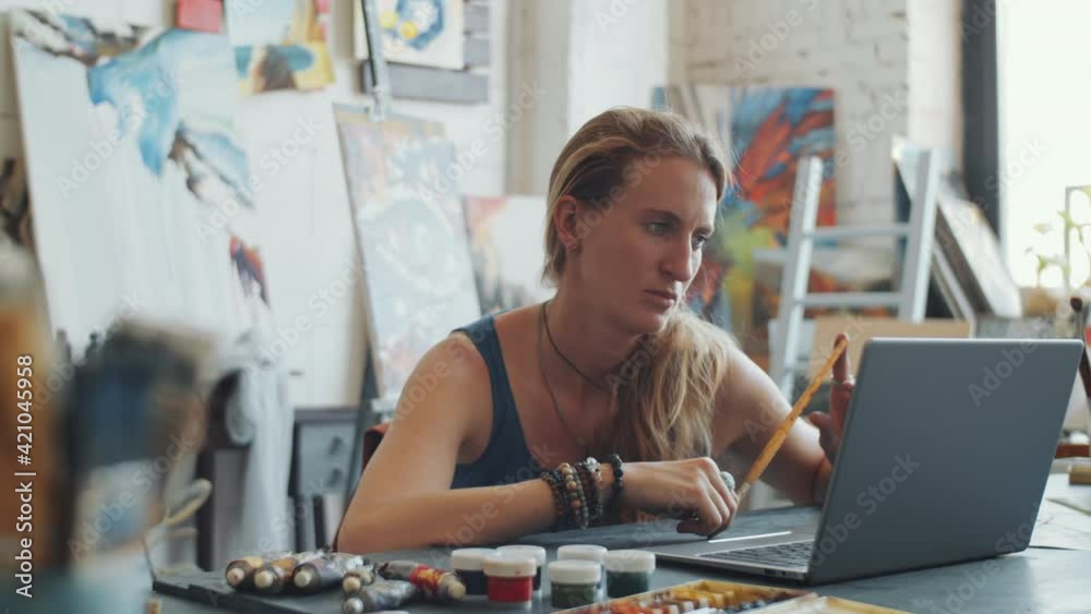 Young cheerful woman waving and speaking on online video call on laptop while sitting at desk in art studio Young cheerful woman waving and speaking on online video call on laptop while sitting at desk in art studio