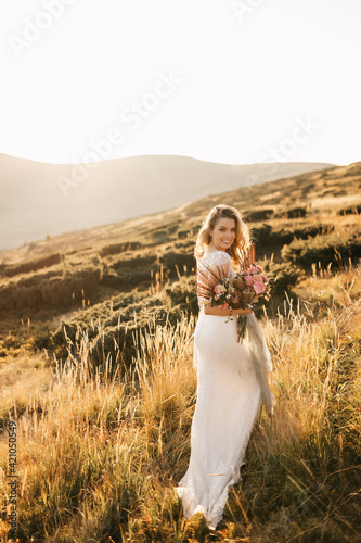 Beautiful bride in a white wedding dress stands posing and smiles in the mountains with a bouquet