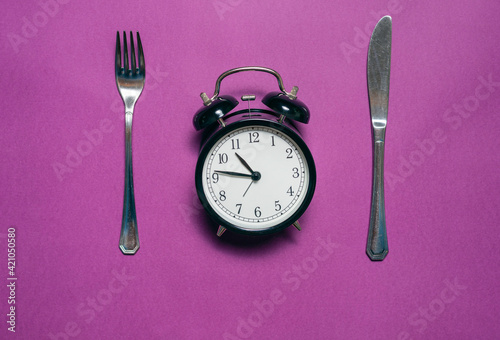  Alarm clock with knife and fork on purple background Conceptual eat up time, lifestyle