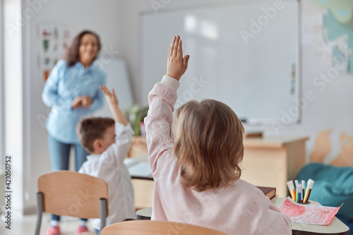 Young happy elementary school pupils at light modern classroom are rising their hands on a lesson