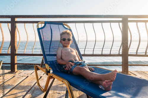 Caucasian funny preschool boy lying on beach chair at the sea. Blonde hair kid in sunglasses sunbathing resting at vacation