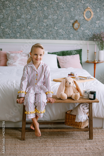 Child girl sitting in pajamas in the bedroom Blonde with blue eyes next to a toy beige plush hare a cup with cocoa and a candle Stylish interior Model portrait