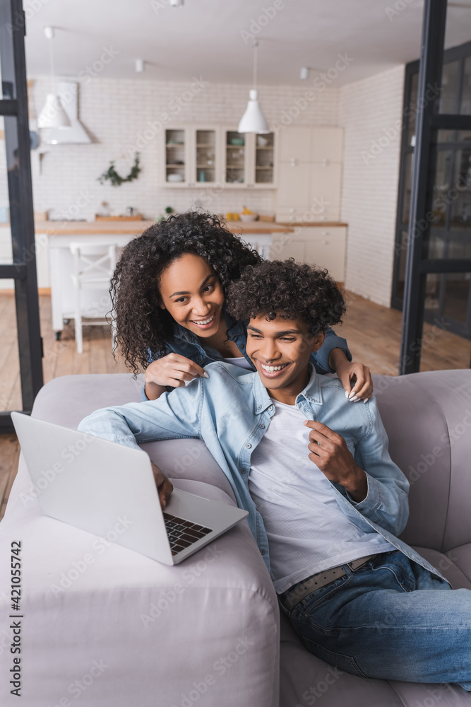 Smiling african american woman hugging boyfriend with laptop in living room