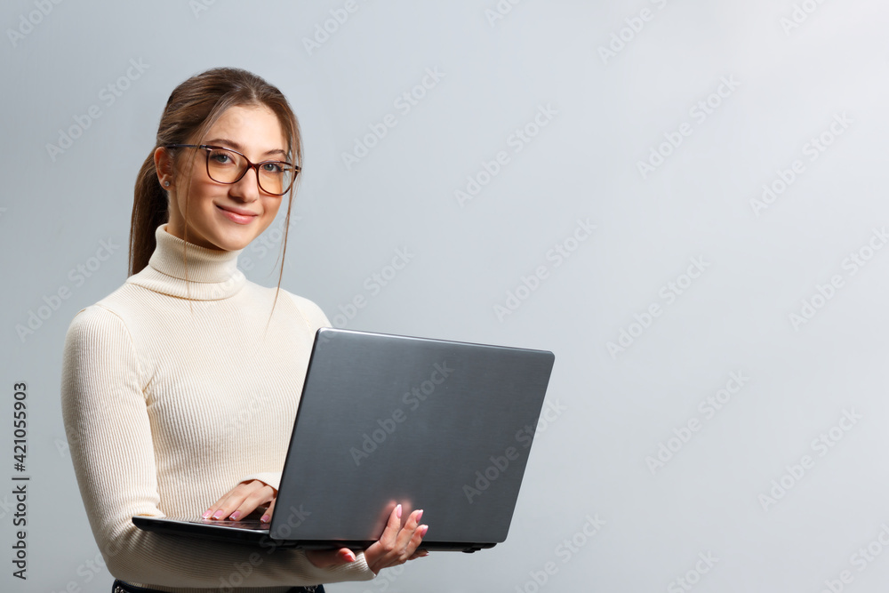 Cute young woman in glasses holds a laptop. Beautiful girl student smiling and looking at the camera on a gray background with copy space. Cute young woman in glasses holds a laptop. Beautiful girl student smiling and looking at the camera on a gray background with copy space.