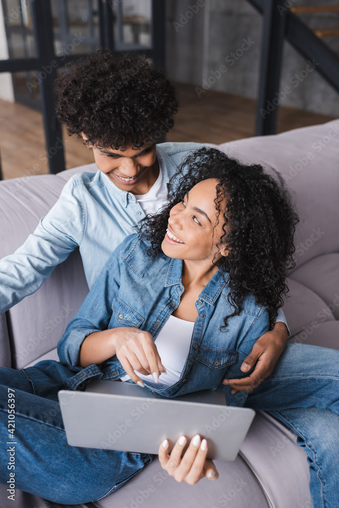 Smiling african american woman holding laptop near boyfriend at home