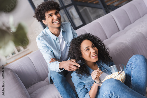 Smiling african american man holding remote controller near girlfriend with popcorn