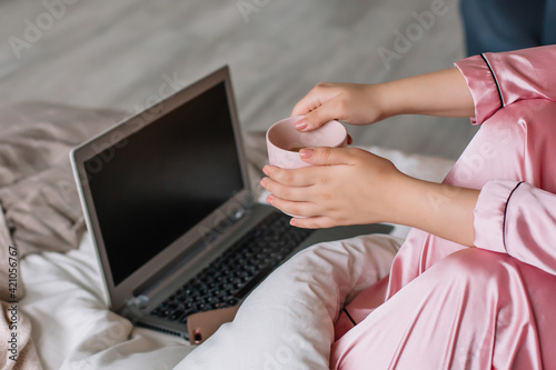 Female hands hold a mug of coffee Pink pajamas on the body Bed in the bedroom with a blanket Laptop and mobile phone lie on the blanket