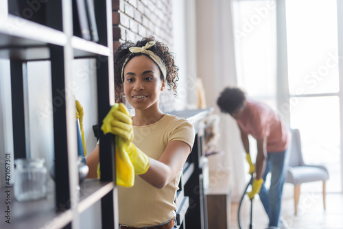 Smiling African American woman cleaning cupboard near boyfriend on blurred background