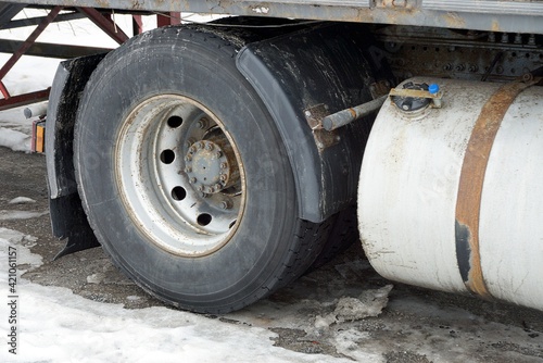 part of an iron white gas tank and one black wheel on a truck stands on gray asphalt and white snow on a winter street