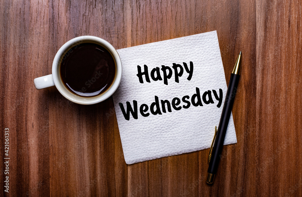On a wooden table next to a white cup of coffee and a pen is a white paper napkin with the words HAPPY WEDNESDAY On a wooden table next to a white cup of coffee and a pen is a white paper napkin with the words HAPPY WEDNESDAY