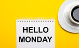 On a bright yellow background, a white cup with coffee and a white notepad with the words HELLO MONDAY