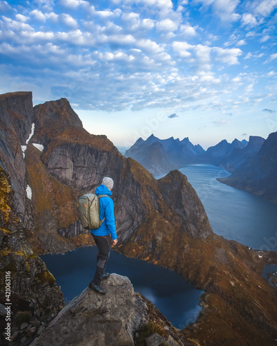 A man with a blue jacket, a hat, and a backpack standing on the top of the mountain, overlooking a beautiful lake and a sea in Lofoten, in Norway during the golden hour.