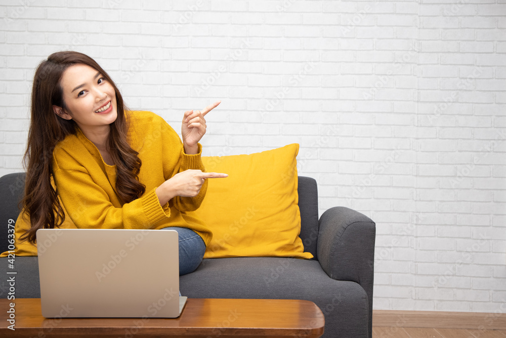 © comzeal - Young elegant beautiful Asian woman smiling and pointing to empty copy space and sitting on sofa with laptop in the living room at home © comzeal - Young elegant beautiful Asian woman smiling and pointing to empty copy space and sitting on sofa with laptop in the living room at home