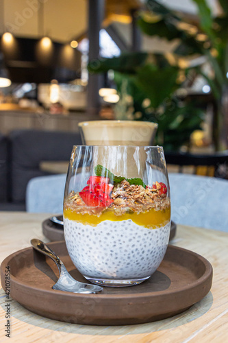 Chia pudding with mango puree, oats flakes and fresh strawberries and cup with cappuccino on wooden table in cafe.