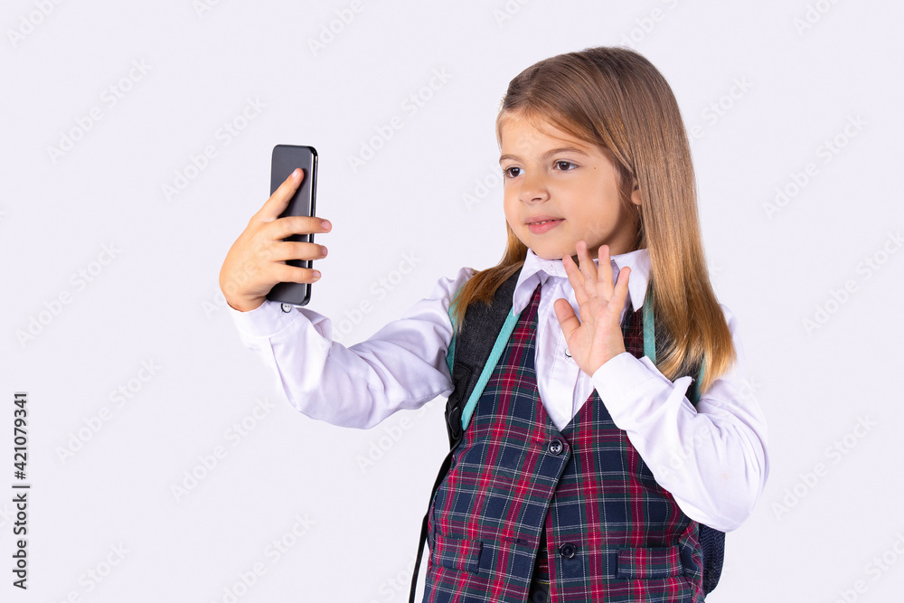 A beautiful preschool girl in uniform greets by video communication on a smartphone. Online training during a pandemic on a white background. A beautiful preschool girl in uniform greets by video communication on a smartphone. Online training during a pandemic on a white background.