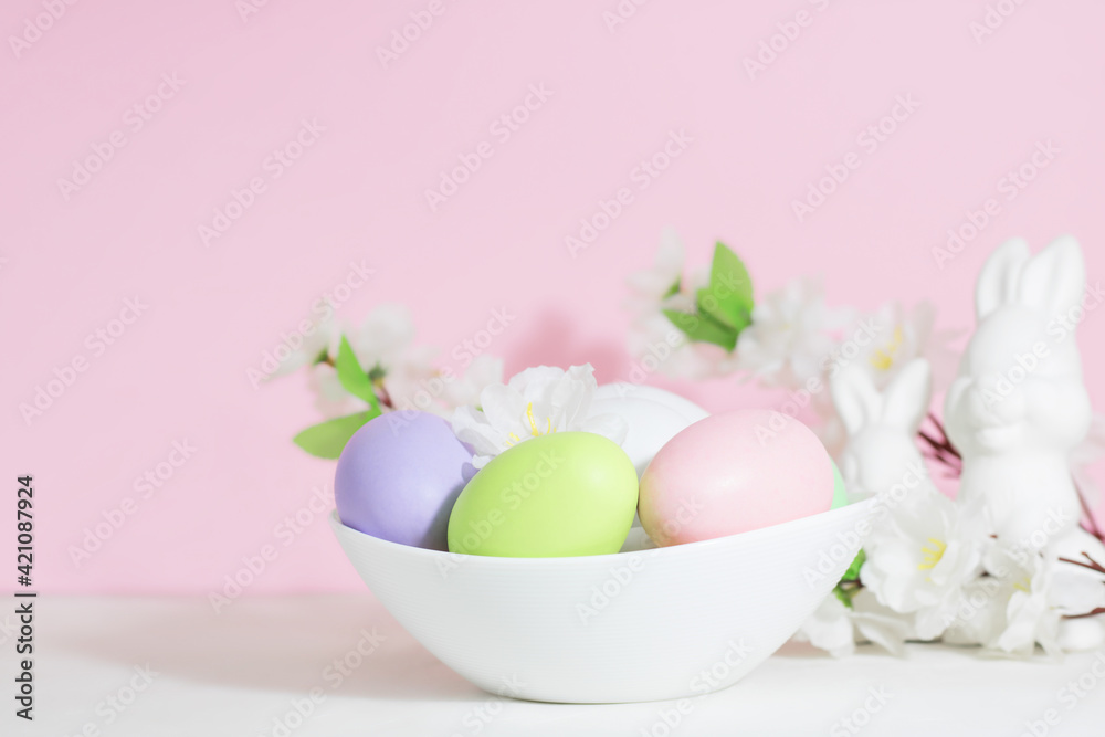 Easter eggs, pink, green and lilac, in a white plate, on a concrete white table, next to bunnies and flowers, on a pink background. Pastel shades.