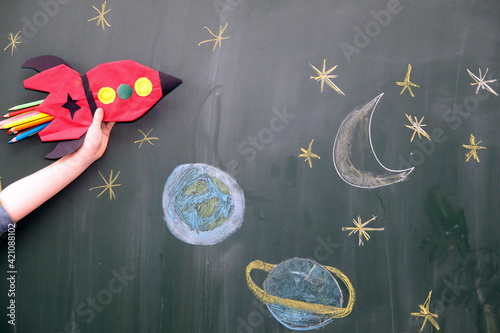 Child hand with handcrafted red rocket and painted planets in front of school blackboard. 