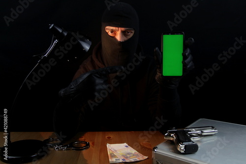 Criminal man wearing balaclava holding smartphone with green screen pointing with finger, over black background.
