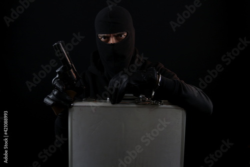 Man with balaclava sitting, holding gun and briefcase attached wrist with handcuffs. Crime concept