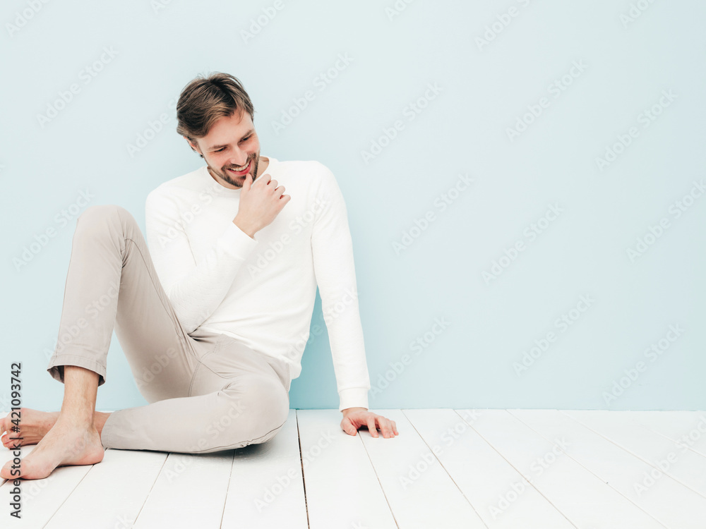 Portrait of handsome smiling hipster lumbersexual businessman model wearing casual white sweater and trousers. Fashion stylish man sitting against light blue wall in studio