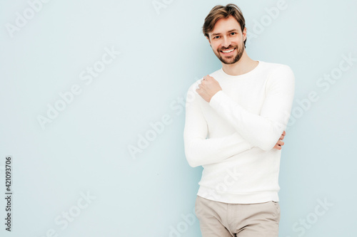 Portrait of handsome smiling hipster lumbersexual businessman model wearing casual white sweater and trousers. Fashion stylish man posing against light blue wall in studio