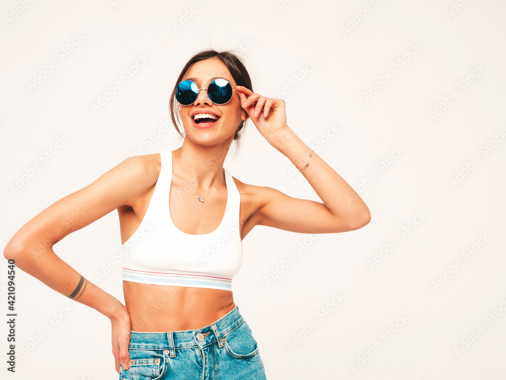 Obraz premium Beautiful smiling woman dressed in white jersey top shirt and jeans. Sexy carefree cheerful model having fun indoors.Adorable and positive female posing on grey background in studio in sunglasses