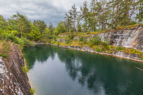Beautiful nature landscape view of rain drops on pond surface. Rocky coast and tall pine trees on pale sky background. Sweden. © Alex